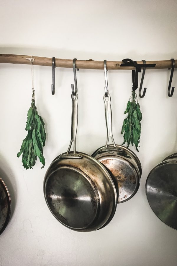 Metal pots and pans and bundles of herbs hang from a stick on a kitchen wall.